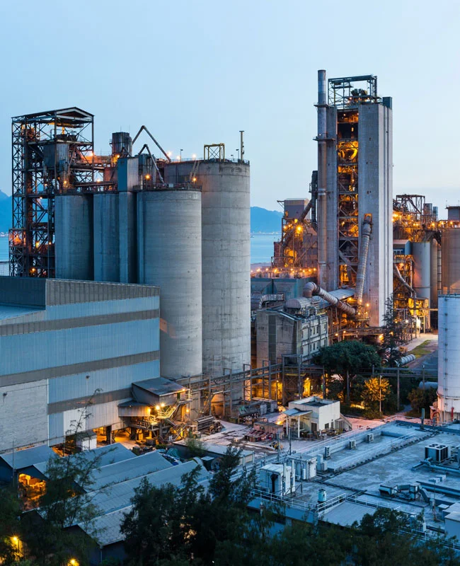 Gran planta de cemento o planta de procesamiento industrial con iluminación nocturna y silos gigantescos cerca del agua.